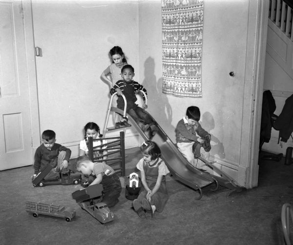 Children playing on and near a slide indoors in the Neighborhood House, 768 West Washington Avenue across from Brittingham Park. Pictured on the slide are: Betty Schuetz and Edward Johnson. Left to right are: Joseph Troig, Jeraldine Stafell, Mary Lou Rubin, Ed Abbott, and Richard Stafell.