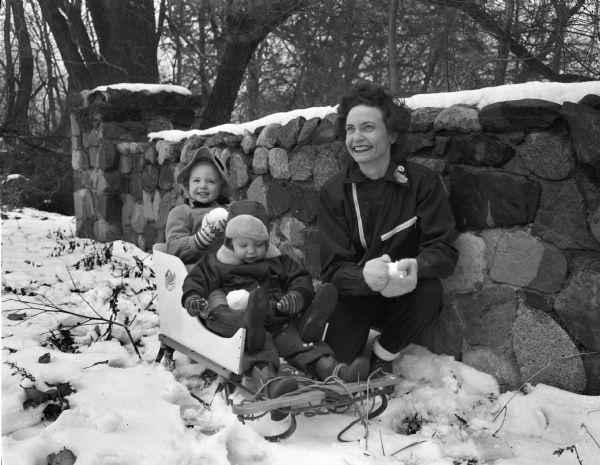 Winter scene with Betty Ann and Arthur G. Field on a sled in the snow with their mother Elizabeth Field kneeling next to them near a stone wall. The family lives at 333 North Randall Avenue.