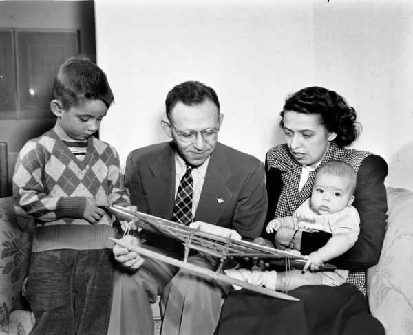 Family portrait of a man, woman, infant, and young boy (wearing argyle sweater), looking at an airplane model.