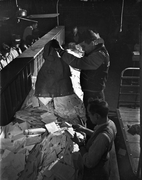 Elevated view of two postal employees sorting mail at the Madison post office, 215 Monona Avenue, at Christmas.  Pictured are clerk Donald Memhard dumping letters onto the facing table where James Kaye is sorting them. Other men are working on the left side of a wooden shelf.