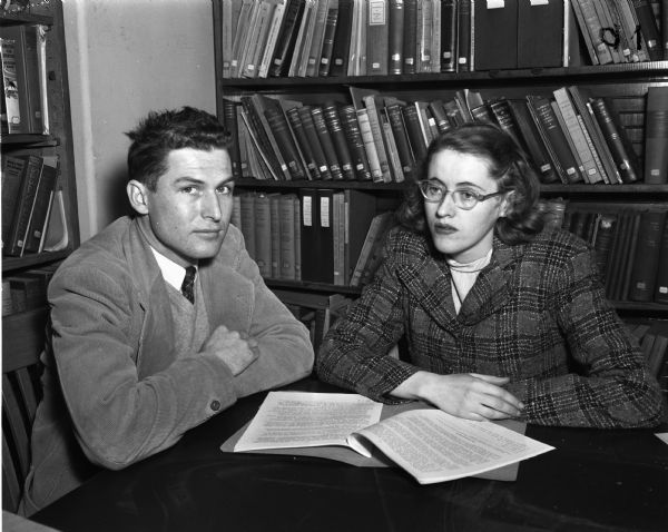 A man and woman seated at a table in front of bookshelves.