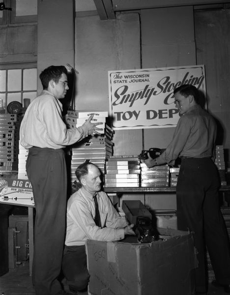 Three workers setting up "Santa's Shop" at the Empty Stocking Club's toy depot at the Madison Community Center, 16-20 East Doty Street. The club is sponsored by the <i>Wisconsin State Journal</i>. Standing left to right are volunteer workers: John Tewes and Bill Doudna, Jr.  Kneeling is Jack Morrell, State Journal employee.
