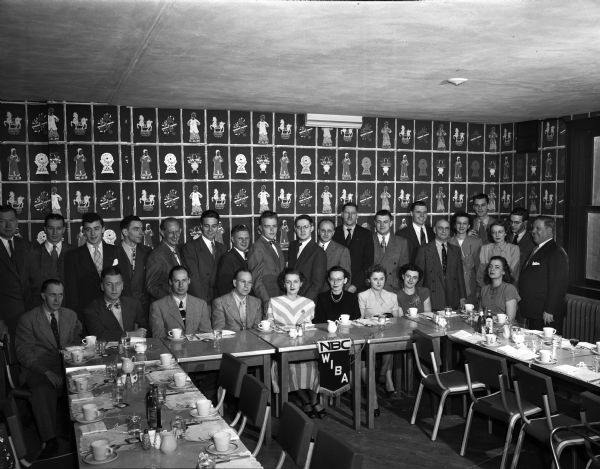 Group portrait at a party for radio station WIBA personnel. People are posing sitting and standing around tables set for dinner in front of a wall with  Asian-themed wallpaper.