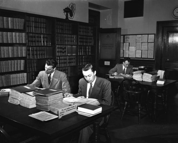 Three men seated at tables using the reference materials in the Legislative Reference Bureau library. Seated at the front table left to right: Wesley Hillery and Anton Jacobson. At the rear table is Victor Hornbostel.
