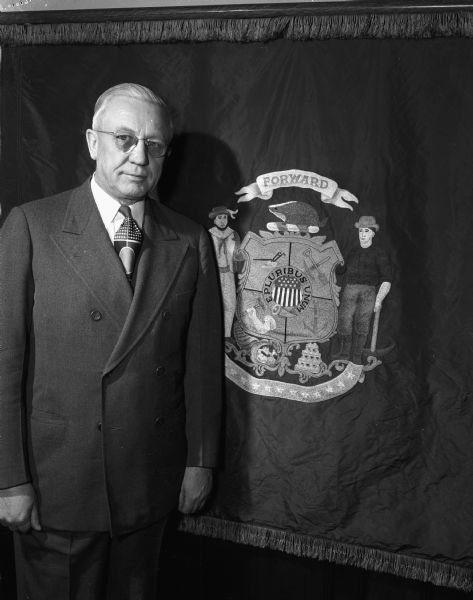 Governor Oscar Rennebohm standing near the state flag of Wisconsin.