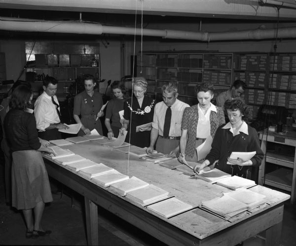 Assembling educational materials for servicemen-students all over the world are the employees of the United States Armed Forces Institute (USAFI), headquartered at 102 North Hamilton Street. Left to Right:  Mary Pfister, George Banker, Robert Corcoran, Madeline Baer, LaVerne Burke, Ruth Herthel, Charles Jacobs, Dorothy Chapin, Arline Sheldon, and at the machine in the back, Russell Olson.