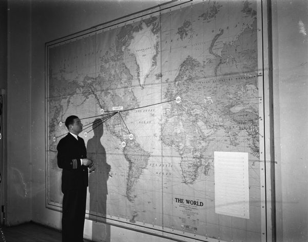 Commander O.W. Goepner standing in front of a map showing overseas armed forces units being served by the United States Armed Forces Institute (USAFI) which provides college level course work to servicemen-students. Seventy colleges and universities are cooperating with USAFI in providing materials and offering credit. USAFI is headquartered at 102 North Hamilton Street.