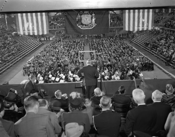 Rear view of Governor Oscar Rennebohm at the speaker's stand on the first balcony in the University of Wisconsin-Madison Field House at a "kick-off" event for the Wisconsin centennial celebration.  Governor Rennebohm is introducing Charles E. Bohlen, counselor to the U.S. State Department and event speaker. The Governor is facing an audience estimated at 3,000. Two large U.S. flags and one large Wisconsin state flag are hanging from above over the speaker's stand.