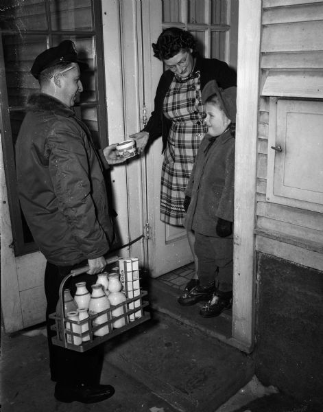 Milk delivery man in work clothes being greeted at the door of a residence by a woman and young girl as he delivers milk and dairy products.  Shown in the photograph is a built-in milk delivery door. Taken for the American Dairy Association.