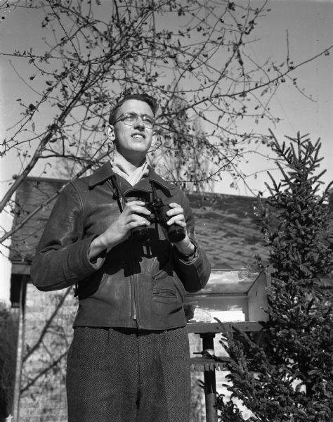 George E. Koehler, son of Mr. & Mrs. Arthur Koehler, 109 Chestnut Street, won a trip to Washington, D.C., for his bird watching and bird census work. He is a senior at West High School and a member of the Four Lakes Drum and Bugle Corps. He is standing in front of a bird feeder holding binoculars, and there is a building in the background.
