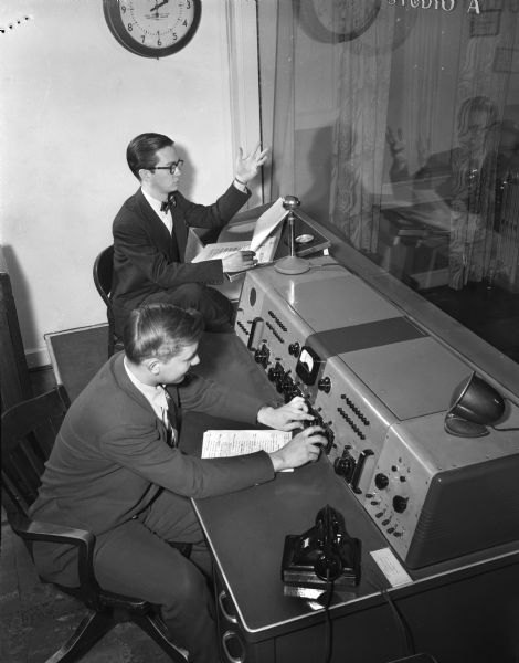 Supervising a WHA radio broadcast are Karl Schmidt, dramatic producer, and Ernest Engberg, studio operator. They are sitting at a desk in front of a window.