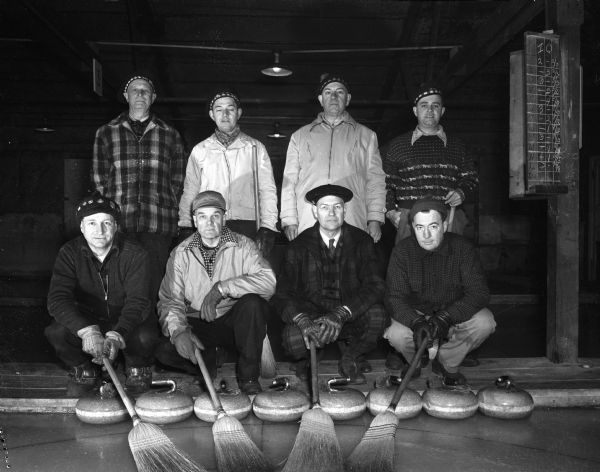 Members of the winning rinks in the intra-club bonspiel, are, standing, the Wellman Rink: left to right, Walter Wellman, Jim Cottrell, Dr. L. F. Fundell, and Bob Wellman; kneeling and holding brooms on the ice are members of the Quilty Rink: left to right, Frances Quilty, John Hill, Gary Larson, and R. J. Peck. The bonspiel was held at the Madison Curling Club at Jones/Burr Field. The men are wearing sweaters, coats and hats. There is a scoreboard on the right.