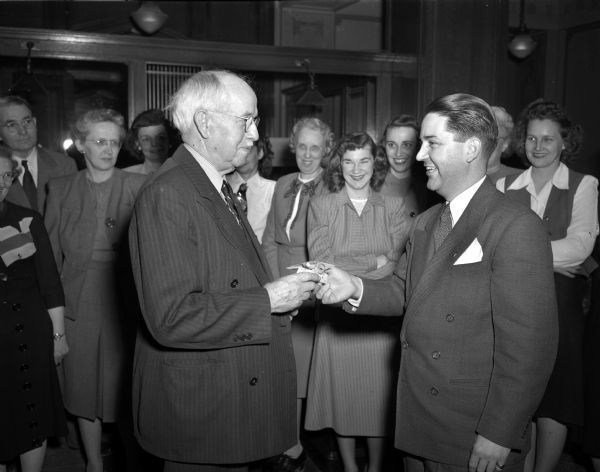 State Treasurer John Sonderegger, on right, presenting a tie pin to his chief accountant, Arthur Pugh, wearing eyeglasses on left, at his retirement. A group of men and women stand looking on in the background.