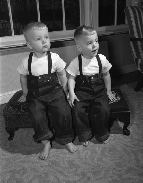 Twin boys Galen Rolf (left) and Mark Rudolf Hasler, sons of Professor and Mrs. Arthur D. Hasler. They are wearing white short-sleeved t-shirts, matching overalls, and are barefoot. They are sitting on footstools near an exterior window.
