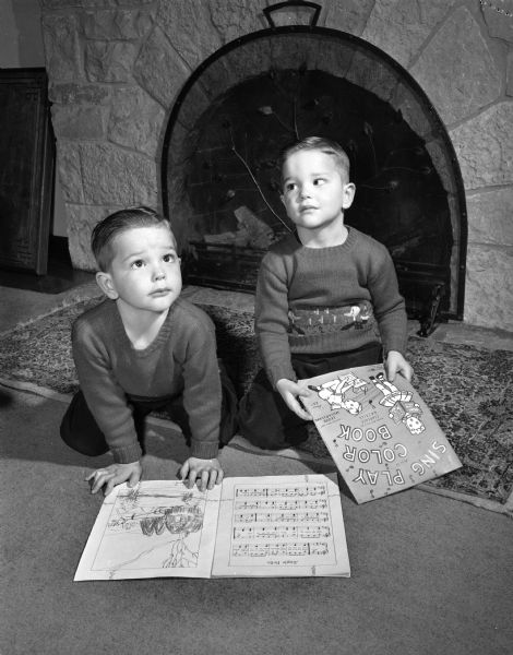 Twin boys Donald (left) and Ronald Jacobson, sons of Mr. and Mrs. Irving Jacobson. They are holding coloring and music books, and are sitting on a rug in front of a fireplace.