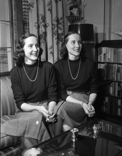Widely known twin sisters in southern Wisconsin, Cecelia (left) and Alma Moore, now Mrs. Walter M. (Cecelia) Mass, Jr., and Mrs. Robert O. (Alma) Bauch. They are sitting on a couch in matching sweaters, skirts, and necklaces.