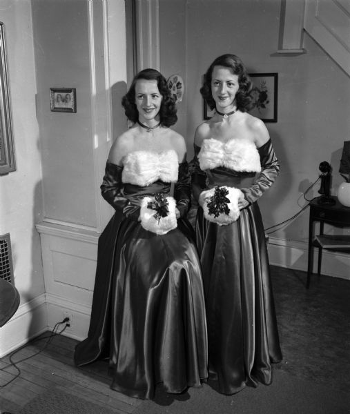 Twin sisters Patricia (left) and Nancy Coughlin in matching formal dresses. They are posing indoors and are wearing matching chokers around their necks and holding muffs decorated with ribbons and holly.
