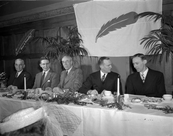 Madison Community Union annual dinner meeting at the Park Hotel. Attending are: left to right, Chief Justice Marvin Rosenberry, Lowell Frautschi, Dr. H.C. Bradley, D.A. Forsberg, and Henry D. McKay. Behind the men on the wall is a banner with a red feather, and to the left is another feather with the words: "Wear It Proudly!"