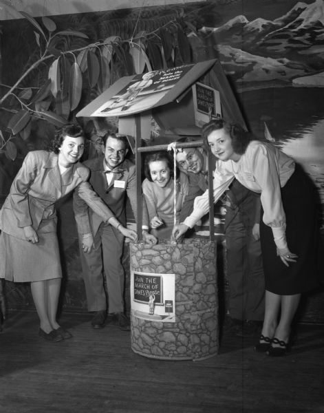 Five members of the Young Adult Club dropping coins into a wishing well during a dance at the Community Center to aid the March of Dimes. Pictured left to right: Lois Pease, Tom Maloney, Gwen Clark, Ben Prochaska, and Rosalyn Gunderson. There are signs and posters that say "Join the March of Dimes Please," and a large plant and a mural are in the background.