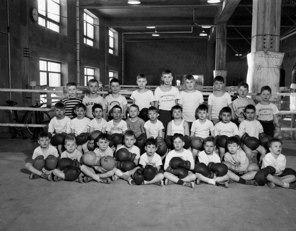 Group portrait of the 28 boys enrolled in the 9 o'clock "Little Badgers" boxing class held at the University of Wisconsin-Madison boxing quarters on Saturday mornings. The boys are wearing shorts and boxing gloves. The boxing ring is in the background, and there is a training bicycle on the left.