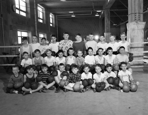 Group portrait of 29 boys enrolled in the 10 o'clock boxing class held at the University of Wisconsin-Madison boxing quarters on Saturday mornings. The boys are wearing shorts and boxing gloves. The boxing ring is in the background, and there is a training bicycle on the left.