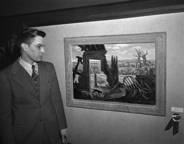 Tom Dietrich, Appleton, juror for the Madison Artists Exhibition, standing next to the painting "Black Cat and Ruins" by Mrs. Fred Logan, winner of the "Award of Excellence."