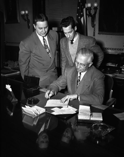 Governor Oscar Rennebohm at his desk signing a proclamation declaring National Security Week, with two men observing behind him.