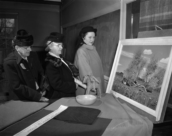 Three women looking at an exhibit at the University of Wisconsin Farm and Home Week, showing room furnishing revolving around rural paintings. Pictured left to right are Mrs. Charles Booth, Blue Mounds; Mrs. Bernard Fetzer, Grimm; and Stasia Lonergan, Portage.