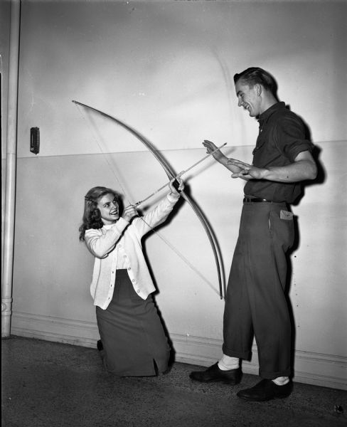 Phyllis Berg plays Cupid with Richard Pigorsch in preparation for a Valentine's Day dance as an example of the various seasonal dances held by different high school groups. Phyllis is kneeling on the floor with a bow and arrow pointed at Richard who is standing.