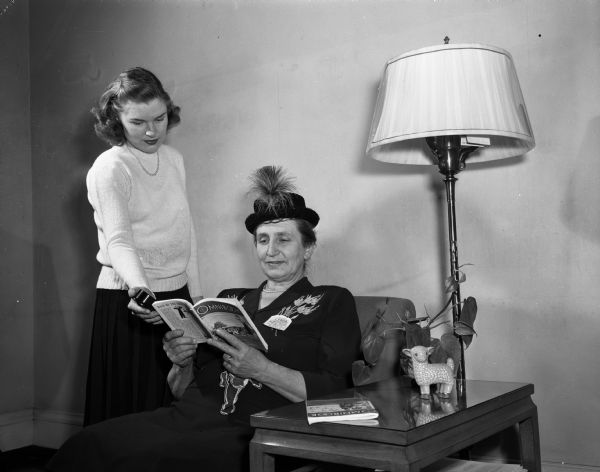 A young woman serving as the photographer's assistant is standing beside a woman sitting in a chair wearing a hat, and a Farm and Home Week name tag. She is reading a magazine called <i>Omnibook</i>. The woman standing is holding a light meter takning a light reading.
