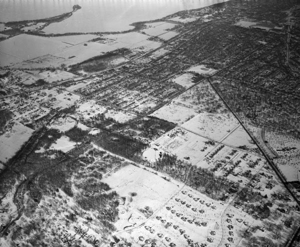 Aerial photograph of Madison's west side looking northeast along Speedway Road. Visible are Forest Hill and Resurrection Cemeteries, West High School, Hoyt Park, Picnic Point, Forest Products Laboratory, and Camp Randall Stadium. Major streets include: Regent Street, University Avenue. There is snow on the ground.