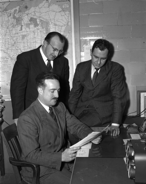 Instructors of the University of Wisconsin Industrial Management Institute staff. Seated at a desk is Associate Professor Russell Moberly, institute director. Standing left to right: instructors Dick Sullivan and Ted Widder, Jr. There is a map of Wisconsin in the background.