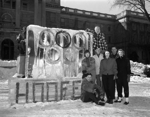 Ice carving of the Olympic rings at the University of Wisconsin Hoofers Club Winter Carnival, with Bascom Hall and the Lincoln Monument in the background. Seven students are pictured next to the ice carving.