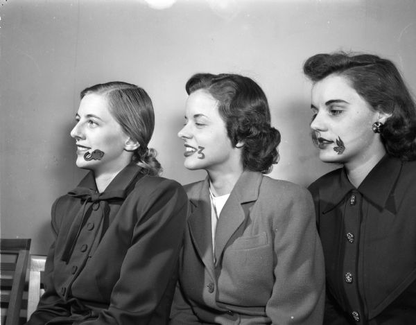 Three University of Wisconsin co-eds sporting "outsize" beauty spots lampooning a fad that is sweeping the country for a stye show sponsored by the University home economics department.  Pictured from the left are: Barbara Savey, Wauwatosa, with a beauty spot in the shape of an elephant head representing the Republican Party; Natalie Bergstrom, Neenah, with a donkey head representing the Democratic Party; and Mariel Kolb, Berlin, with the number 3 representing Third political parties.
