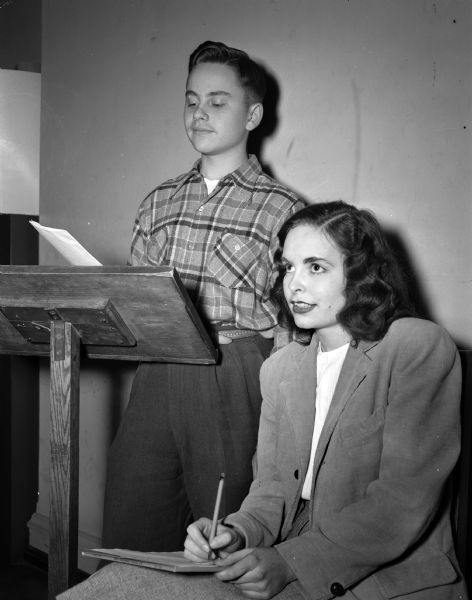 Sherwyn Woods, standing at a podium, and Joanne Conlin, sitting with pad of paper and pencil. They are members of the negative debate team of Wisconsin High School.