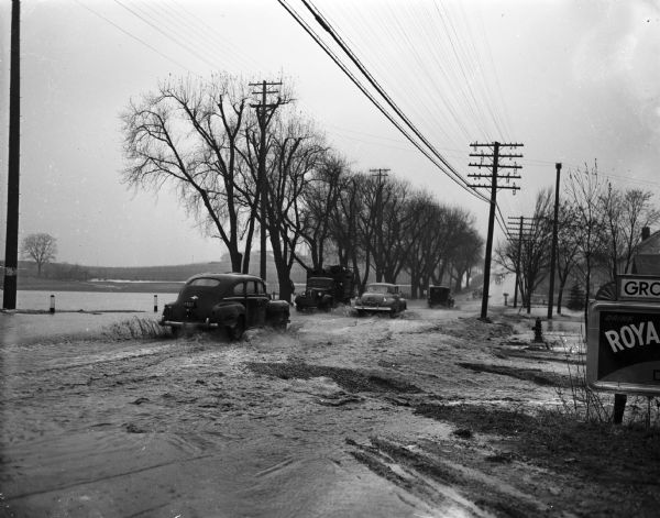 Flood at the University of Wisconsin-Madison East Hill Trailer Camp, located at 451 North Midvale Boulevard, south of University Avenue.  Looking west, showing several vehicles traveling on University Avenue at the north end of the trailer camp. Shown at right is part of a sign advertising Gustav Kieswalter, Groceries and Meats.
