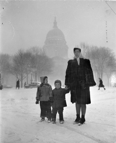 A winter scene of the Capitol Square during the year's record snowstorm.  Pictured are Mrs. Francis (Mayme) Harrison, 304 West Main Street, with her two children, Donna Mae and Dean. The Wisconsin State Capitol is in the background, partially obscured by the heavy snow.