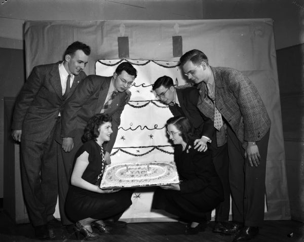 Young Adult Club of Madison celebrates its second anniversary with a dance. Six young adults pose by a cake, in front of a painted backdrop also decorated with a cake.