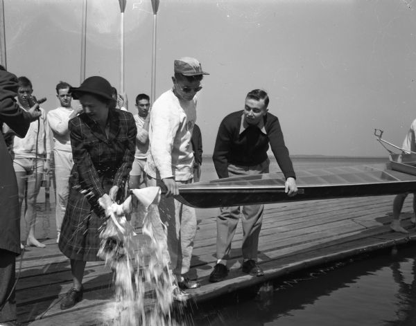 Wisconsin's new racing shell, the <i>Mendota</i>, receives the champagne bottle from sponsor Mrs. Harry (Mary) Stuhldreher in the christening ceremony on the University of Wisconsin armory pier.  The boat, made of pressed plywood, is believed to be the first shell constructed in the middle-west. Coxswain Carlyle Fay is grasping the prow of the <i>Mendota</i>.