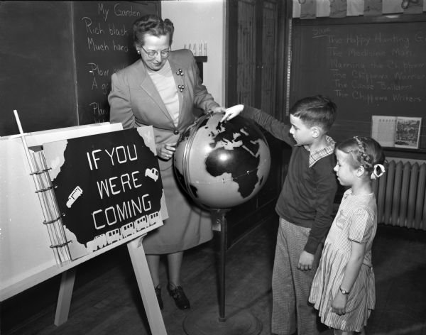 Mrs. Everett (Helen) Holterman, Randall School teacher, and student Nancy Hoff, look on as classmate Lonny Koff points out the country of Norway on a world globe.