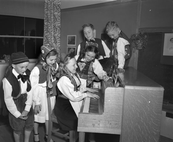 Six students from Marquette School representing early Wisconsin settlers in Norwegian ethnic costumes performing in the school pageant celebrating Wisconsin's centennial. Gathered around the piano singing a Norwegian song are, left to right: Robert Strand, Louise Revell, Annette Larson, Sue Strand, David Bickel, and Douglas Lindquist.