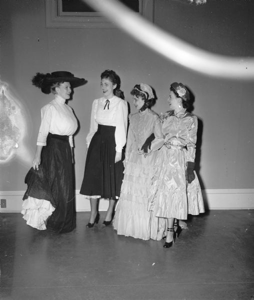 Four University of Wisconsin Alpha Chi Omega sorority sisters modeling clothes from "then and now". Pictured from left to right are: Larinice Ballam in a 1905 Gibson Girl outfit; Marily Coon in a 1948 version of a Gibson Girl outfit; Mrs. James L. Severson in a 1880's hoop-skirted gown; Mrs. Wade Plater in a 1948 full-skirted frock.