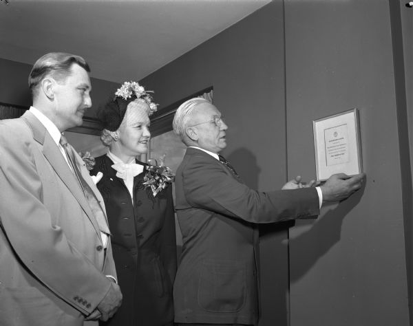 Governor Oscar Rennebohm hanging a dedication plaque on the wall of the Governor's suite in the new Edgewater Hotel, 666 Wisconsin Avenue. Shown looking on are Dr. A.A. Quisling, president of the Edgewater Corporation (left), and Mrs. Oscar (Mary) Rennebohm (center).