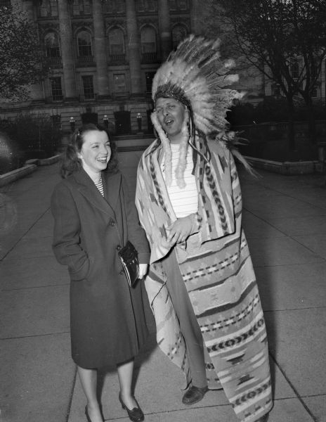 Crawford County historical pageant performers came to Madison to announce the Crawford County historical pageant and Villa Louis opening at Prairie du Chien. Jerry Dunbar, dressed as an Indian, is telling Mary Ward, 110 South Henry Street, about the event. In the background is the Wisconsin State Capitol.