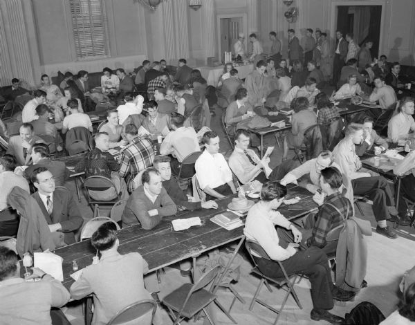University of Wisconsin students referred to as the "Lunchbox Brigade" seated at tables in the Great Hall of the Memorial Union on the campus eating their home-packed lunches. In the background is a service counter set up by the Union for the sale of beverages, soups and desserts.