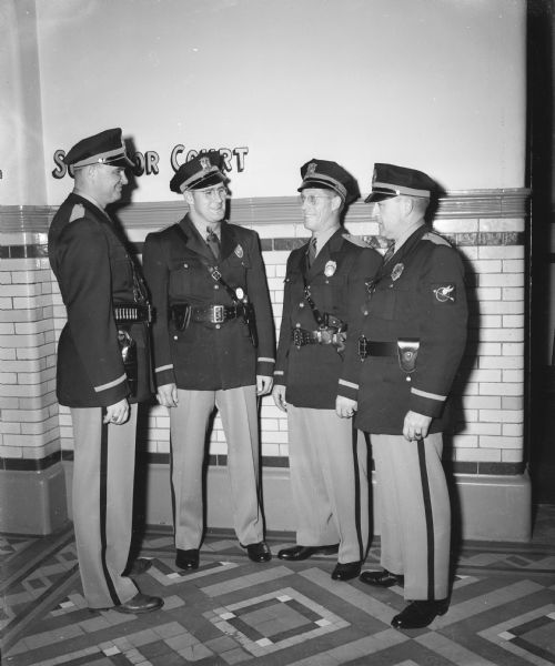 Four Dane County traffic police pictured in their new navy blue and grey uniforms. Left to right, Officers Charles O'Brien, Don Harless, Emil Schmale, and Lt. Arden C. Pope. They are standing in a building with a sign on the wall that says, in part: "S...or Court." Probably Superior Court.