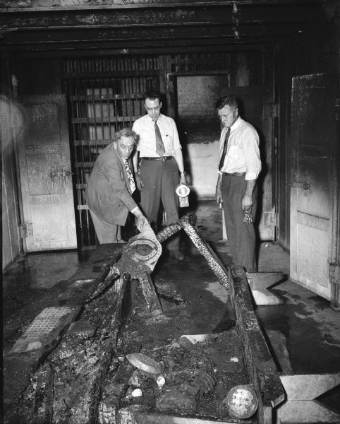 Public officials inspecting the damage and debris from a fatal fire in the Dane County Jail. The fire was started by two inmates, one of whom died in the escape attempt. Left to right: Sheriff Edward A. Fischer, Deputy Reynold Abrahams, and Jailer Neal Smithback.