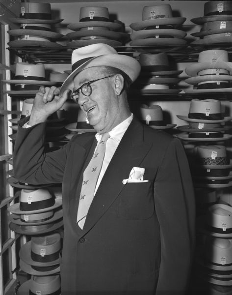Joseph L. "Roundy" Coughlin, columnist for the <i>Wisconsin State Journal</i>, trying on a straw hat. In the background are racks of hats.