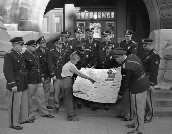 Dane County traffic police with Jimmy Christensen pointing to his plans for a soap box racer. The traffic police are sponsoring Jimmy in the Soap Box Derby. The photograph was taken standing on the steps of the Dane County Court House.