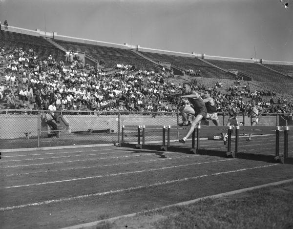 Preliminary hurdle race at the Big Nine Track Meet in Camp Randall Stadium. A crowd watches from the stands.
